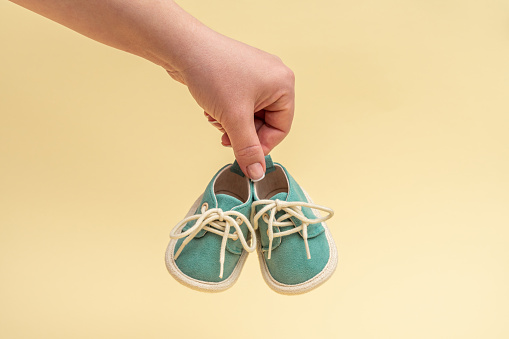A pair of small shoes of a newborn baby in a female hand on a yellow background. Newborn concept.