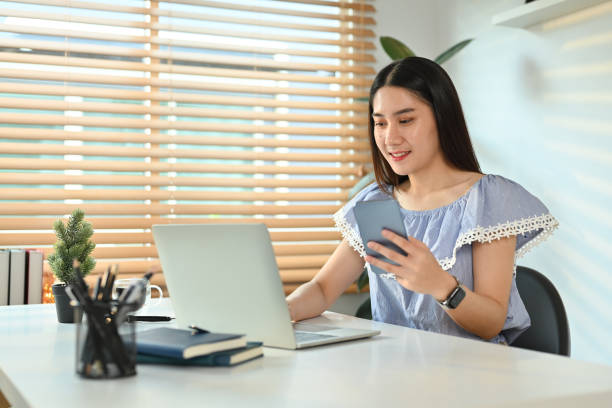 istock Charming female small business entrepreneur checking information on smart phone and using laptop at workstation. 1478018519