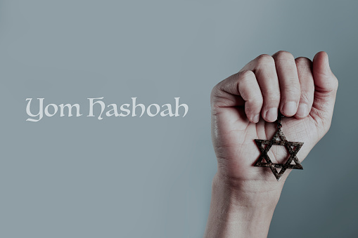 the text Yom Hashoah and the hand of a man holding an old and rusty pendant in the shape of the star of David on a gray background