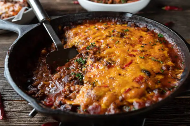Homemade tex mex dish with a delicious and spicy beans stew. Cooked with ground beef, baked beans, kidney beans, bell peppers, onions, tomatoes, garlic and herbs. Gratinated with a tasty cheddar cheese, chili topping. Served in a cast iron pan on wooden table.