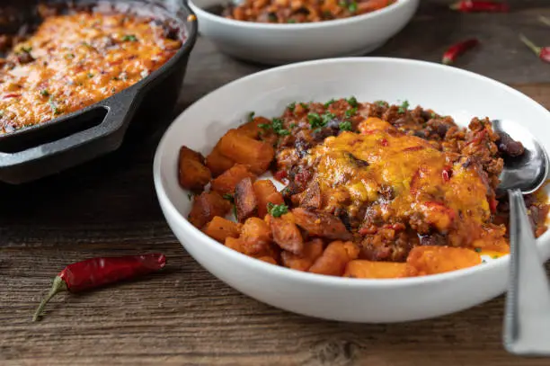 Homemade chili con carne with delicious chili cheddar cheese topping. Baked in a cast iron pan and served with roasted sweet potatoes on wooden table background. Closeup, front view