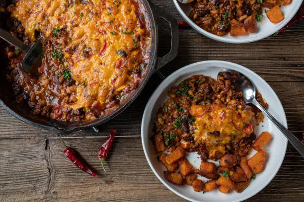 Delicious and savory tex mex dish with a spicy bean stew. Cooked with kidney beans, baked beans, ground beef and vegetable. Gratinated with cheddar cheese, chili topping. Served ready to eat in a cast iron pan on wooden table from above