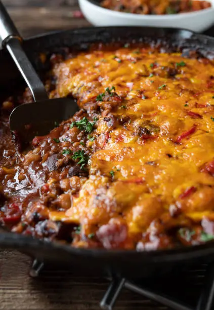 Homemade tex mex food with a delicious and spicy bean stew with ground beef, vegetables and cheddar cheese topping. Served hot and ready to eat in a rustic cast iron pan on wooden table. Closeup