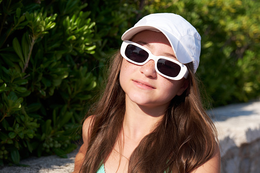 Portrait of a young beautiful girl with freckles in a white cap and sunglasses on the beach on a sunny summer day.