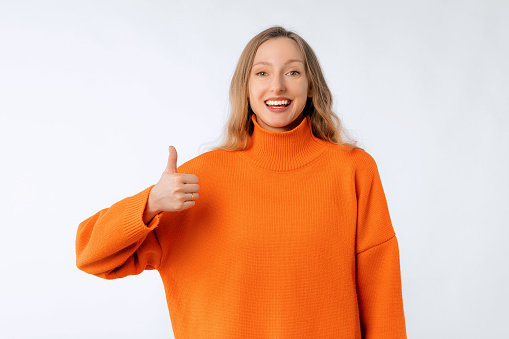 Lovely happy smiling young woman shows thumb up, recommend and approve something good, wears soft orange sweater, standing over neutral studio background