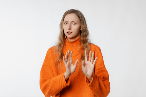 Young woman with hands raised displeased and fearful doing disgust face because aversion reaction, stands in trendy knitwear orange sweater against neutral studio background