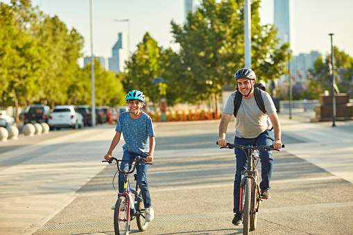 Full length front view of 45 year old man and 11 year old boy wearing t-shirts and jeans, safety helmets, and grinning as they ride through sunny esplanade.