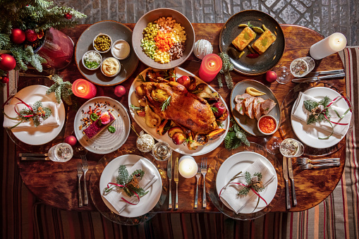 Festive table setting with fried chicken, pear, rosemary, apples, bacon, mustard, red sauce, spruce branches, olivier salad, mayonnaise, gorka, onion, mayonnaise, sour cream, burning candles, cutlery, champagne glasses and Christmas toys