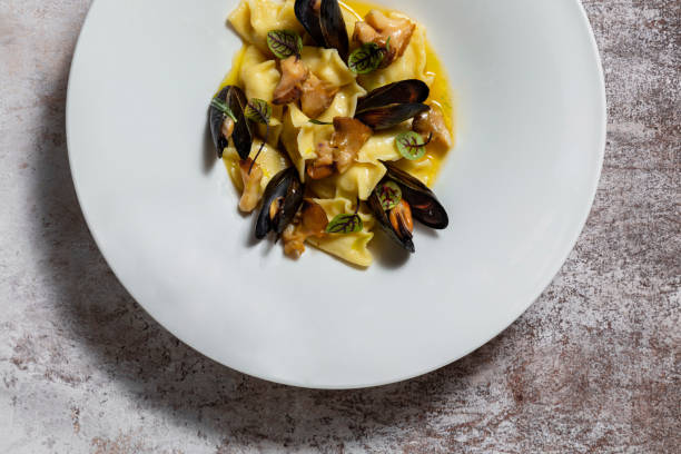Vareniki stuffed with sturgeon fillet. Mussels and rapanas, lettuce leaves and wine sauce are poured on top. The food lies in a white, round, ceramic plate. The plate stands on a dark, coffee-colored fabric background. vareniki relleno de filete de esturión. mejillones y rapanas, hojas de lechuga y salsa de vino se vierten encima. la comida se encuentra en un plato blanco, redondo y de cerámica. el plato se encuentra sobre un fondo de tela oscura de color café.