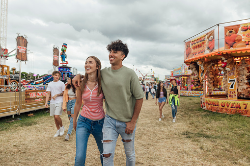 Group of teenagers enjoying a funfair in Newcastle, North East of England. They are walking through a field filled with stalls and rides, laughing and talking together. Two of them have their arms around each other while they walk.