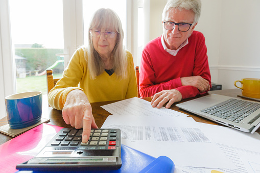A senior couple check their finances at home while surrounded by documents, folders, laptop and a calculator. They both appear slightly worried and anxious.