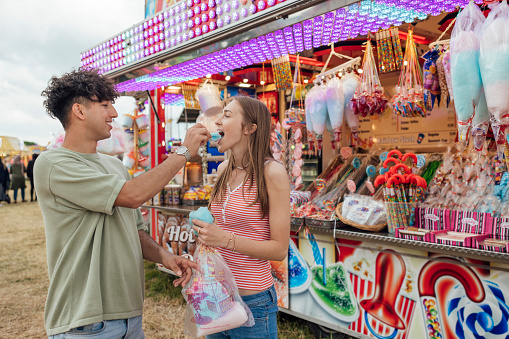 Teenage couple enjoying a funfair in Newcastle, North East of England. They are eating candy floss they have purchases from a food vendor, laughing together while the boyfriend feeds his girlfriend.