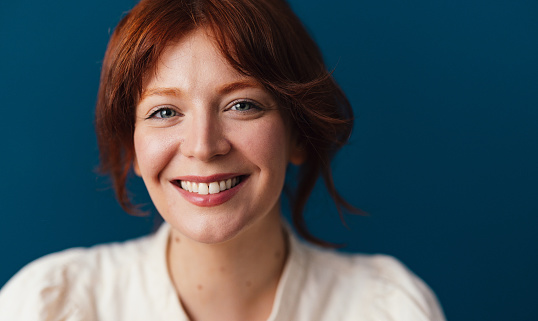 Happy woman with red hair looking at camera while sitting in front of the blue background.