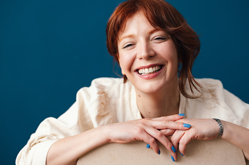 Happy woman with red hair looking at  camera while sitting on a chair in front of the blue background.