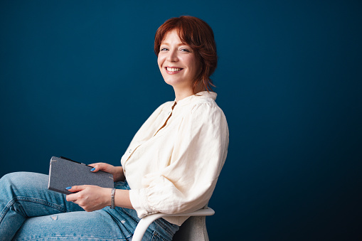 Smiling woman with red hair holding notebook and looking at the camera while sitting on a chair in front of the blue background.