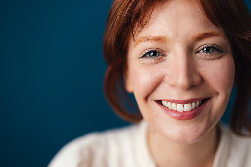 Close up photo of a happy woman with red hair looking at camera while sitting in front of the blue background.