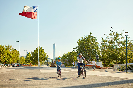 Full length view of mature man and 11 year old boy in casual weekend clothing and safety helmets riding bikes with Chilean flag blowing in background.