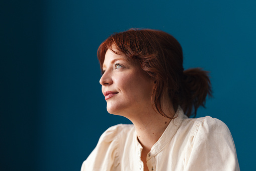 Low angle view of a smiling woman with red hair looking away while sitting in front of the blue background.