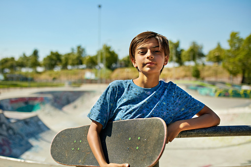 Waist-up front view of 11 year old wearing casual clothing, leaning against railing at sunny urban skate park, and looking at camera.