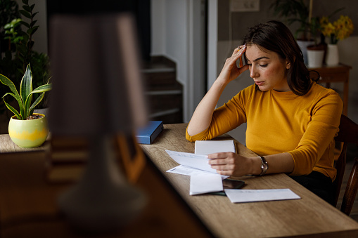 Selective focus shot of worried young woman sitting at her home office desk, head in hand, sorting out her monthly bills and expenses and struggling with finances.