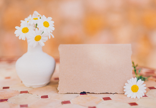 Cute close up of a small vase with white daisies and an empty recycled paper card with a decorative border as a mockup for your Easter, spring or summer text message in front of a mediterranean stone wall. Creative color editing. Very soft and selective focus. Part of a series.