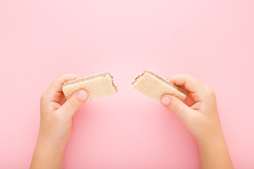 Little girl hands holding broken wafer pieces on light pink table background. Pastel color. Sweet snack. Two pieces. Closeup. Point of view shot. Top down view.