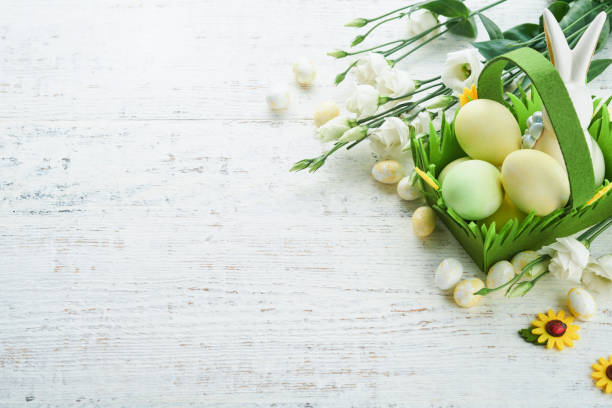 istock Happy Easter. Easter eggs and rabbit in green basket on white old wooden table with white and yellow roses. Spring Happy Easter holiday card.  Easter background with copy space. Top view. 1478085318