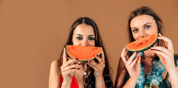 two smiling cheerful caucasian girls with slice of watermelon in front the face on a brown background
