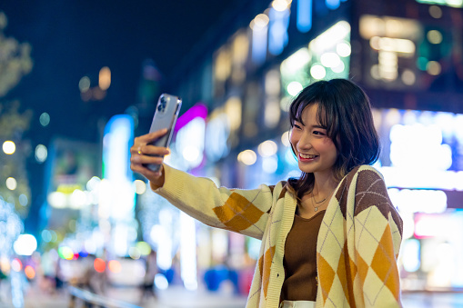 Young Asian woman using mobile phone taking selfie with urban cityscape and street lights at night. Attractive girl enjoy and fun outdoor nightlife travel city with using portable device technology.