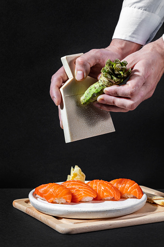 Fresh wasabi root. Male hands hold a special grater for Wasabi Japanese horseradish root in one hand and the root itself in the other. Under the hands lies a board with a brush for cleaning and a light, round, ceramic plate, on which Nigiri with salmon lie. The board lies on a black paper background.