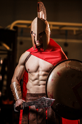 Male portrait of sports man indoors who exercising gym in the gym. Lifestyle photography with athletic people who doing fitness and professional sport. Person using spartan costume with shield, helmet and sword. Sparta warrior.