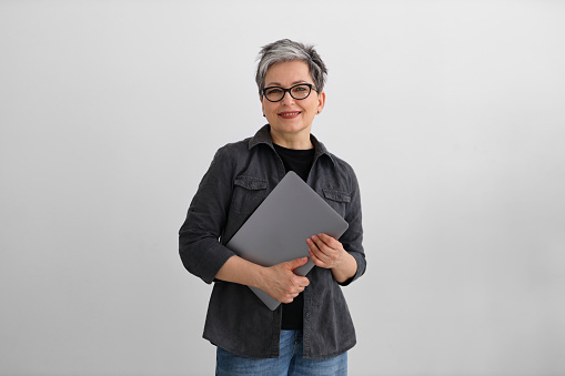 Una programadora adulta vestida de negro sonríe sobre un fondo gris, sosteniendo una computadora portátil en sus manos. photo