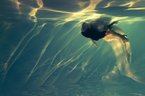 Underwater shoot of beautiful woman in white flying transparent dress swimming in water through sunbeams. Fantasy mermaid against water surface background with rays of lights.
