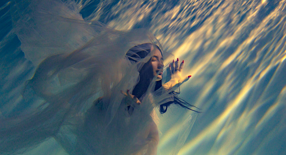 Underwater shoot of beautiful woman in white flying transparent dress swimming in water through sunbeams. Fantasy mermaid against water surface background with rays of lights. Closeup.