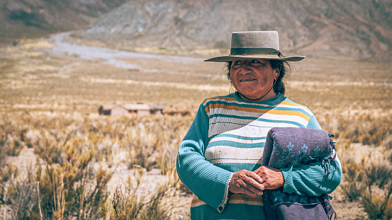 Andean woman with her house on the edge of the mountain in the background. January 2022, Salta, Argentina.