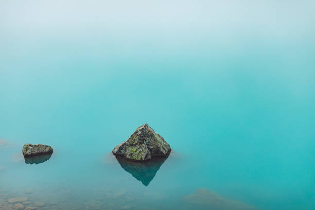 Serene lake with rocks in the foreground and distant mountains under a cloudy blue sky at dusk.