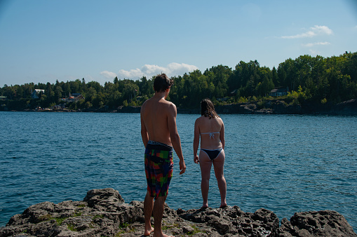A man and a woman in bathing suits stand on the rocky shoreline of the lake during a sunny day in the summer