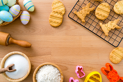 Overhead viev of ingredients and utensils used for preparing Easter cookies at home arranged all around the border of a wooden background making a frame and leaving useful copy space. High resolution 42Mp studio digital capture taken with SONY A7rII and Zeiss Batis 40mm F2.0 CF lens.