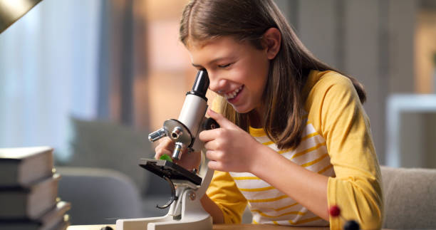 Science student, microscope and girl at desk with homework, education and studying chemistry or physics. Learning, knowledge and analysis, happy child at home with school project and study with smile