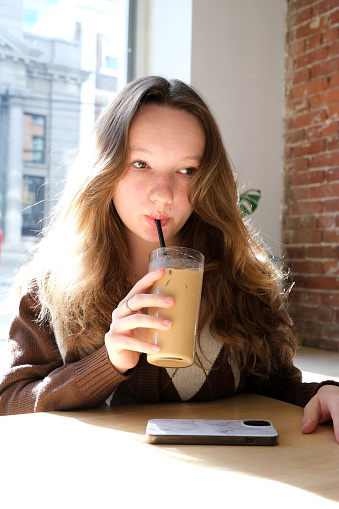 girl in a brown vest sits on a brick background and drinks iced coffee a cold drink rests relaxes thinks licks her lips a teenager in thought black jeans calm pastime waiting for friends loneliness