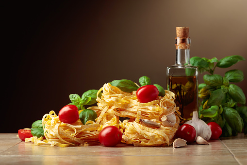 Homemade raw pasta with olive oil, tomato, garlic, and basil on a brown background. Copy space.