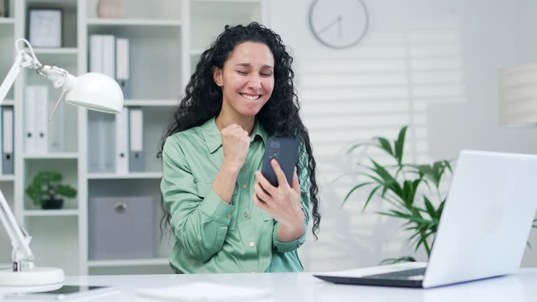 Overjoyed young curly brunette businesswoman extremely happy read message on Smartphone phone at workplace