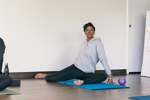 A beautiful African American woman sits on a yoga mat and smiles while doing warm-up stretches before exercising at the gym.