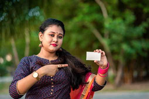 Shot of a cheerful young woman holding a blank business card outside at park with green trees in background.