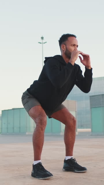 VERTICAL VIDEO: Young bearded male athlete doing workout, squats on the waterfront
