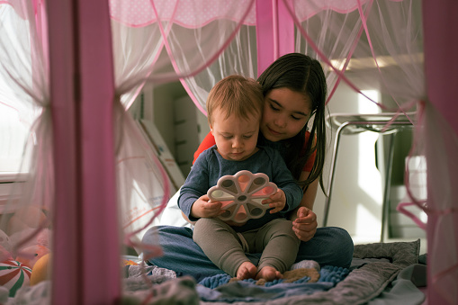 A cute Eurasian toddler boy sits on his sister's lap in a pink castle tent set up in their playroom and plays with a toy.