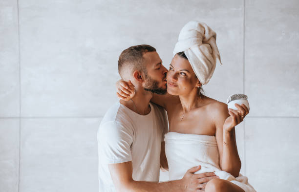 joven barbudo besando a su esposa en una toalla envuelta alrededor de su cabeza en el baño. la mujer hispana morena bronceada sostiene la crema para el cuidado de la piel satisfecha después del spa en el descanso de vacaciones en el hotel. vacaciones, am - two parent family naked men couple fotografías e imágenes de stock