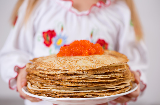 A girl in a Ukrainian embroidered dress is holding pancakes with red caviar on a light background