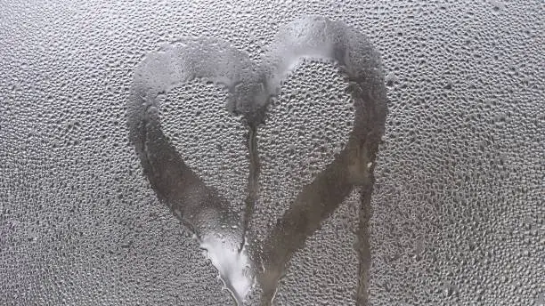 Drawing of a heart on the sweaty glass of the window. Drops run down the glass.