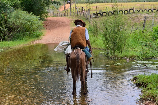 Canudos, Bahia, Brazil - February 1,2023 : A lone mature man rides his horse across the Rio Prieta carrying a bag of groceries